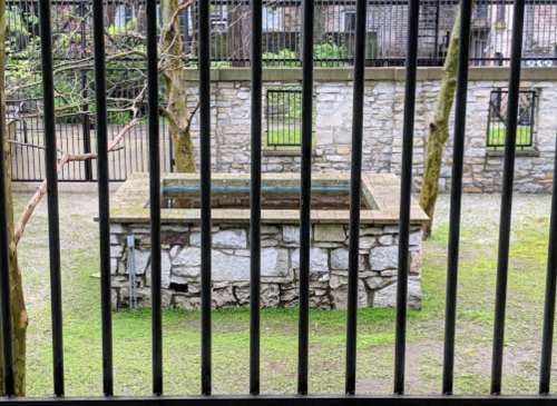 A stone well surrounded by grass, viewed through vertical metal bars of a fence.