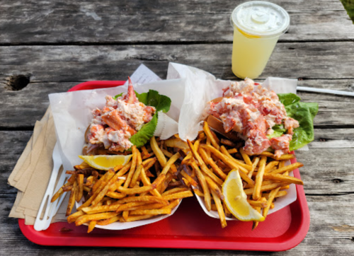 Two lobster rolls topped with lobster salad on a tray, accompanied by crispy fries and a lemon wedge, with a drink in the background.