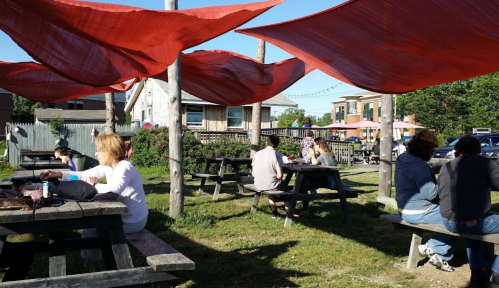 People sitting at picnic tables under red canopies in a sunny outdoor space. Green grass and buildings in the background.