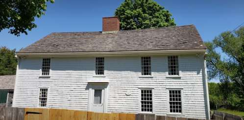 A white, two-story house with a sloped roof and a brick chimney, surrounded by trees and a wooden fence.