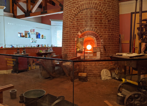A brick furnace glows orange in a workshop, surrounded by tools and materials for glassblowing.