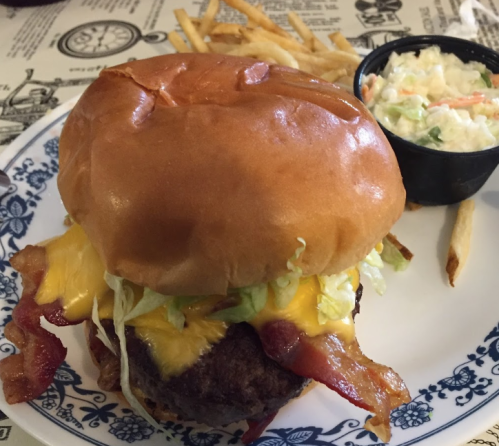 A juicy cheeseburger topped with bacon and lettuce, served with fries and a side of coleslaw on a decorative plate.