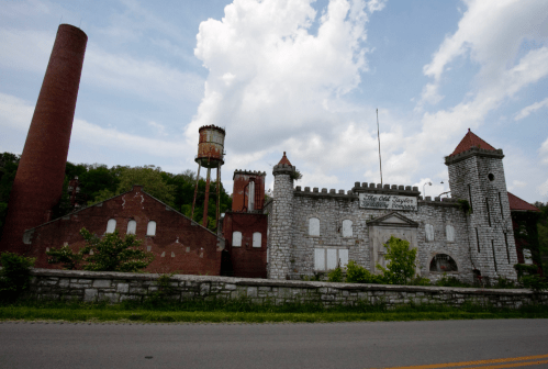 Abandoned stone castle-like building with a tall chimney and water tower, set against a cloudy sky.