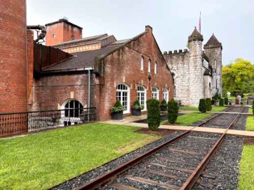 Historic brick buildings with a stone tower, surrounded by green grass and old railway tracks.