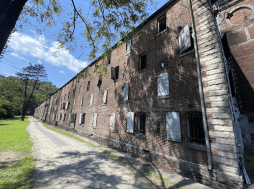Abandoned brick building with boarded windows, surrounded by greenery and a gravel path under a blue sky.