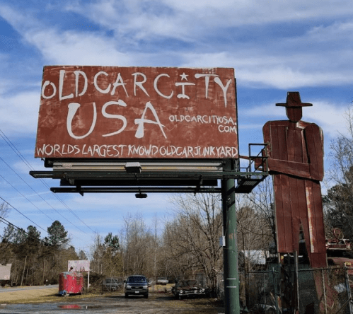 Billboard for Old Car City USA, featuring a large metal figure and advertising the world's largest known old car junkyard.