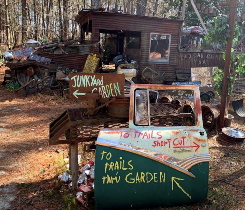 A rustic scene with a signpost pointing to trails and a garden, surrounded by old vehicles and a cabin in the woods.
