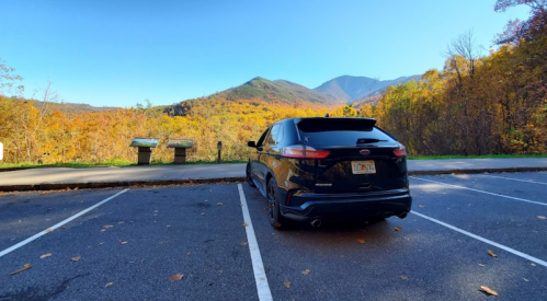 A black car parked in a lot with a scenic view of mountains and autumn foliage in the background.