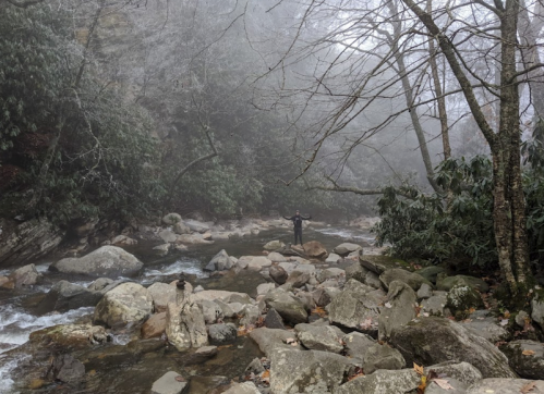 A misty river scene with a person standing on rocks, surrounded by trees and fog.