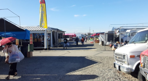 A bustling outdoor market with vendors, colorful tents, and shoppers walking along a gravel path under a clear blue sky.