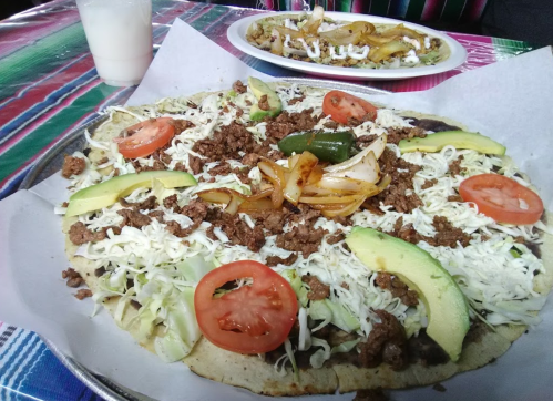 A large plate of taco pizza topped with ground meat, lettuce, tomatoes, avocado, jalapeños, and grilled onions.