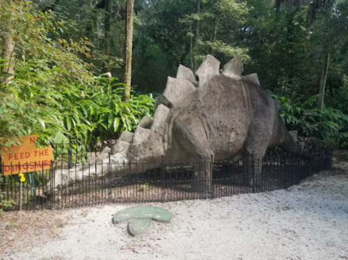 A large dinosaur statue surrounded by greenery, with a sign that says "Do Not Feed the Dinosaurs."