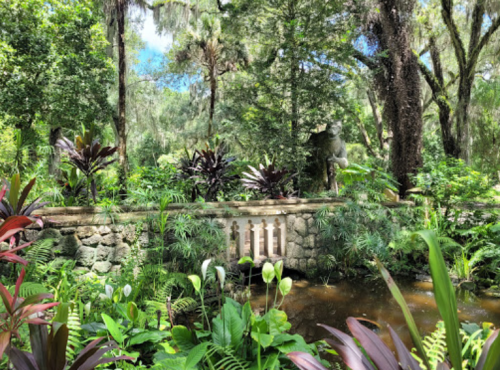 Lush garden scene with a stone bridge, vibrant plants, and a serene pond surrounded by trees.