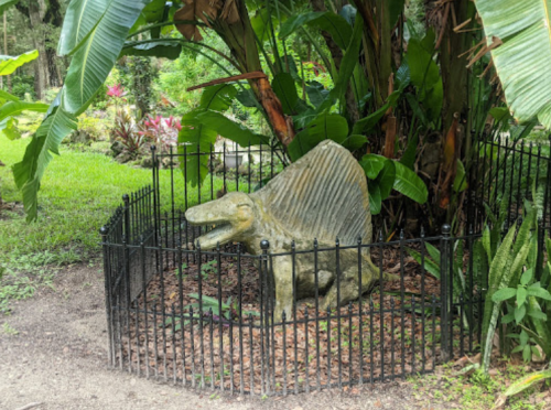 A stone sculpture of a prehistoric creature surrounded by a black fence, set in a lush garden with tropical plants.