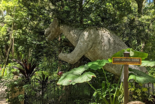 A large statue of a giant ground sloth among lush greenery, with a sign nearby identifying it.