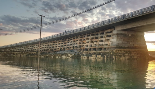 A bridge over calm water, with a textured stone base and power lines in the background under a cloudy sky.