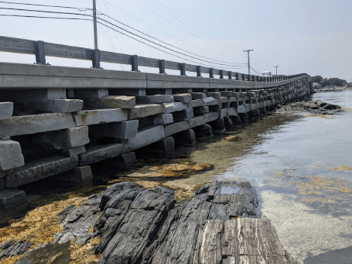 A long bridge extends over rocky shoreline, with clear water and seaweed visible beneath. Power lines run alongside.