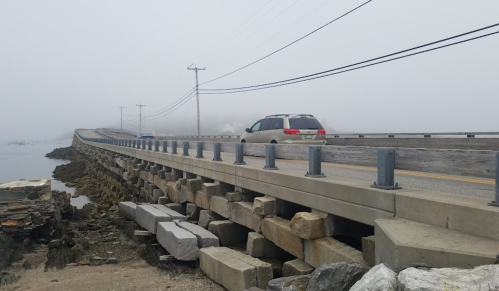 A foggy scene of a bridge over water, with a car driving and rocky supports visible along the side.