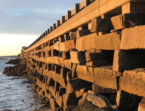 A stone pier extends into the water, illuminated by warm sunset light, with a clear sky in the background.