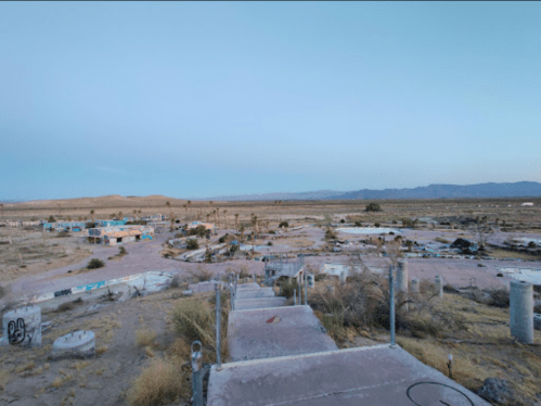 A desolate landscape with remnants of buildings and scattered vegetation under a clear sky. Mountains in the distance.