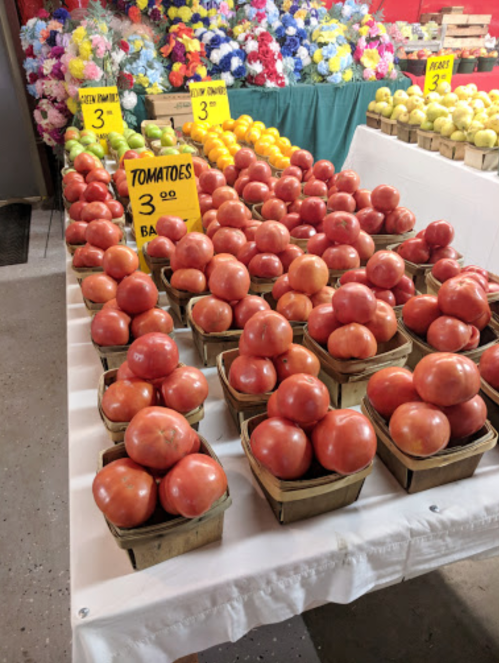 A display of fresh tomatoes in baskets at a market, with colorful flowers in the background and price signs visible.