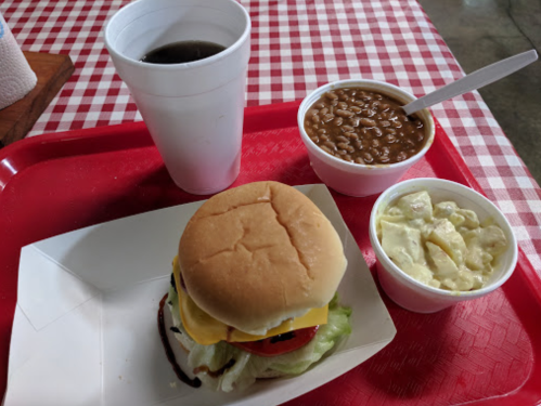 A tray with a burger, macaroni salad, baked beans, and a drink on a red checkered tablecloth.