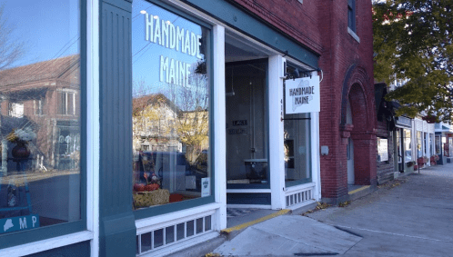 A storefront labeled "Handmade Maine" with large windows, showcasing a quaint street scene in a small town.