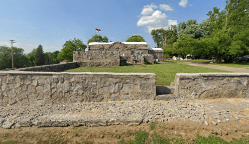 A stone fort with a flag, surrounded by green grass and trees, under a clear blue sky.