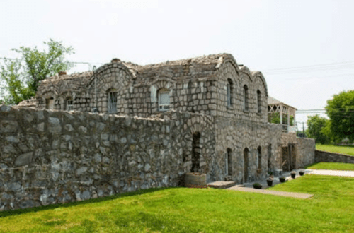 Stone building with arched windows, partially in ruins, surrounded by grass and trees on a sunny day.