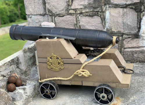 A decorative model cannon on wheels, made of wood, with a black barrel and gold emblem, resting on a stone surface.