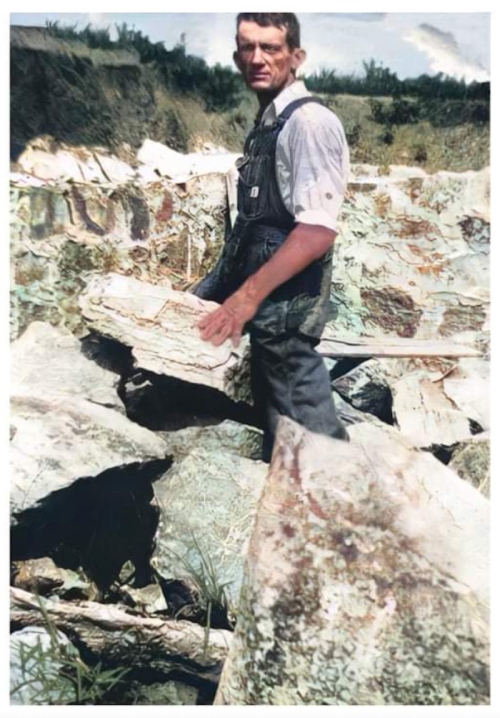 A man in overalls stands among large rocks, holding a piece of stone in a rugged outdoor setting.