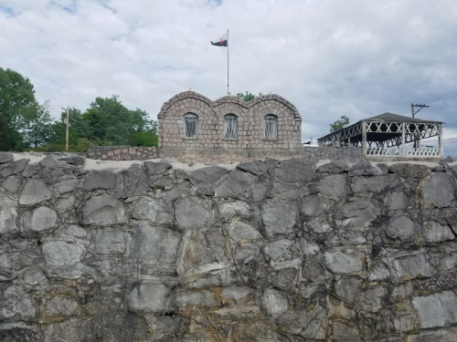 A stone building with arched windows sits atop a rocky wall, with a flag flying above and trees in the background.
