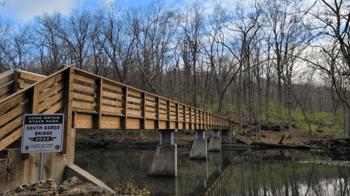 A wooden bridge spans a calm river in a wooded area, with bare trees and green foliage in the background.