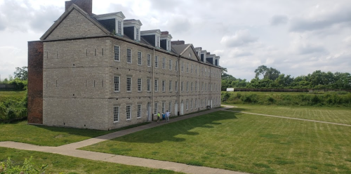 Historic brick building with multiple windows, set in a grassy area under a cloudy sky.
