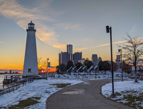 A lighthouse stands by a snowy path, with a city skyline and sunset in the background.