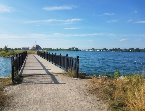 A wooden pier extends into a calm lake, with greenery and a distant building under a clear blue sky.