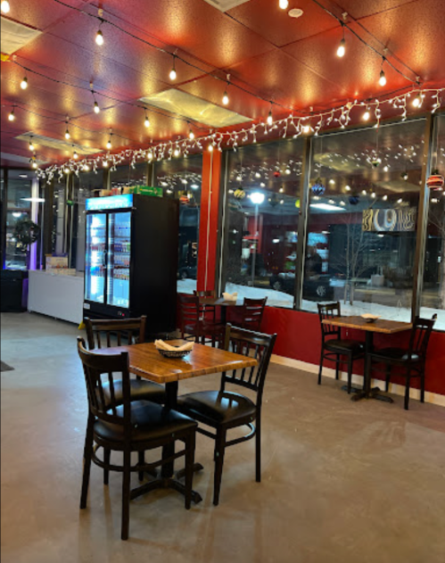 Cozy restaurant interior with string lights, tables, and a drink fridge, featuring large windows and a warm atmosphere.
