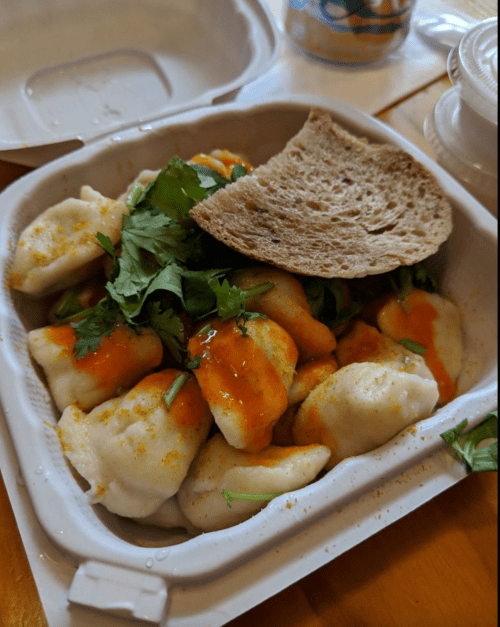 A takeout container filled with dumplings topped with cilantro and sauce, accompanied by a piece of bread.