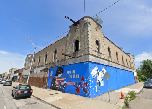 A corner building with a colorful mural featuring a winged figure and the text "KICK OUT THE JAMS." Clear blue sky above.