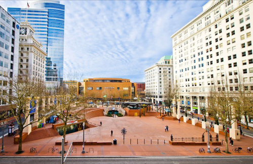 A wide view of a city plaza with trees, buildings, and people walking under a cloudy sky.