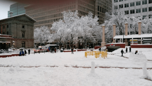 Snow-covered park with people playing, trees frosted white, and buildings in the background.