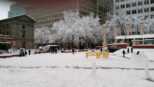 Snow-covered park with people playing, trees frosted white, and buildings in the background.