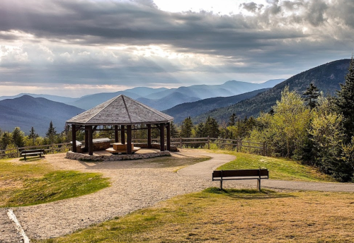 Scenic view of a gazebo on a hill, surrounded by mountains and trees under a cloudy sky.