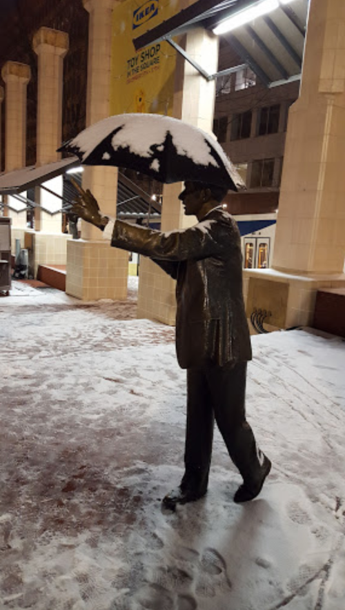 A snow-covered statue of a man holding an umbrella, standing in a snowy urban setting.