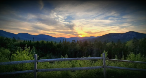 A scenic view of mountains at sunset, with a wooden fence in the foreground and lush greenery below.
