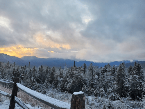 A snowy landscape at sunrise, with mountains in the background and a wooden fence in the foreground.