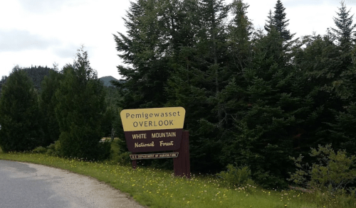 Sign for Pemigewasset Overlook in White Mountain National Forest, surrounded by lush green trees and a gravel road.