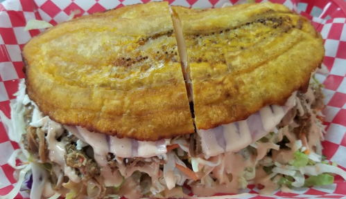 A split plantain sandwich filled with shredded meat, lettuce, and drizzled with sauce, served in a red-checkered basket.