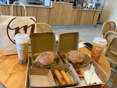 Two boxes of burgers with sesame seed buns, spring rolls, and drinks on a wooden table in a café setting.