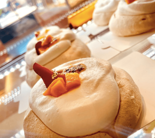 Close-up of meringue desserts topped with cream and fruit pieces, displayed in a glass case.